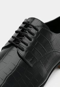 Chaussure habillée en cuir noir avec une texture brillante embossée façon croco et six paires de lacets noirs. Elle présente un bout arrondi et des accents cousus.