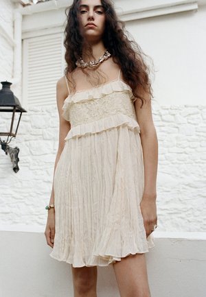 Jeune femme aux longs cheveux foncés portant une robe crème sans manches en dentelle et à volants, debout contre un mur de pierre blanc en extérieur.