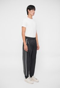 Pantalones deportivos negros con franjas laterales blancas, cintura elástica y bolsillos con cremallera, combinados con una camiseta blanca básica y zapatillas blancas.