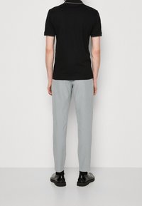 Homme portant un polo noir, un pantalon gris clair, des chaussettes noires et des chaussures habillées noires, debout face à un mur blanc.