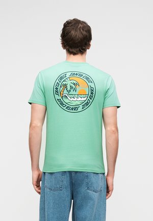 Férfi menta zöld pólóban, amelyen egy Santa Cruz feliratú kör alakú tengerparti naplemente és pálmafa grafikája látható, kék farmerrel párosítva, háttal állva.