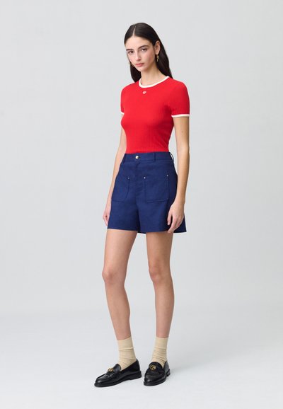 Top corto de manga corta ribeteado en rojo con un borde blanco, combinado con pantalones cortos azul marino que tienen dos bolsillos frontales. Unos mocasines negros y calcetines beige completan el atuendo.