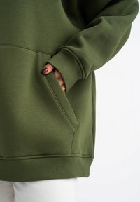 Olijfgroene sweatshirt met een kangoeroezak, ribgebreide manchetten en onderrand. Zachte textuur met een relaxte pasvorm, met aandacht voor het detail van de voorkantzak.