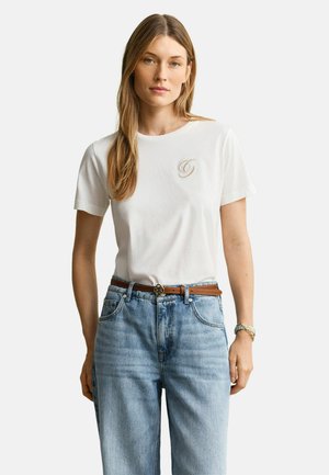 Vrouw met lang haar, gekleed in een wit shirt met korte mouwen en een klein logo op de borst, lichtblauwe spijkerbroek, bruine riem en een polshorloge.