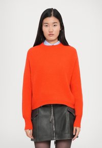 Maglione a coste di un brillante arancione con maniche lunghe, abbinato a una gonna in pelle nera con zip centrale e tasche laterali, indossato sopra una camicia azzurro chiaro.