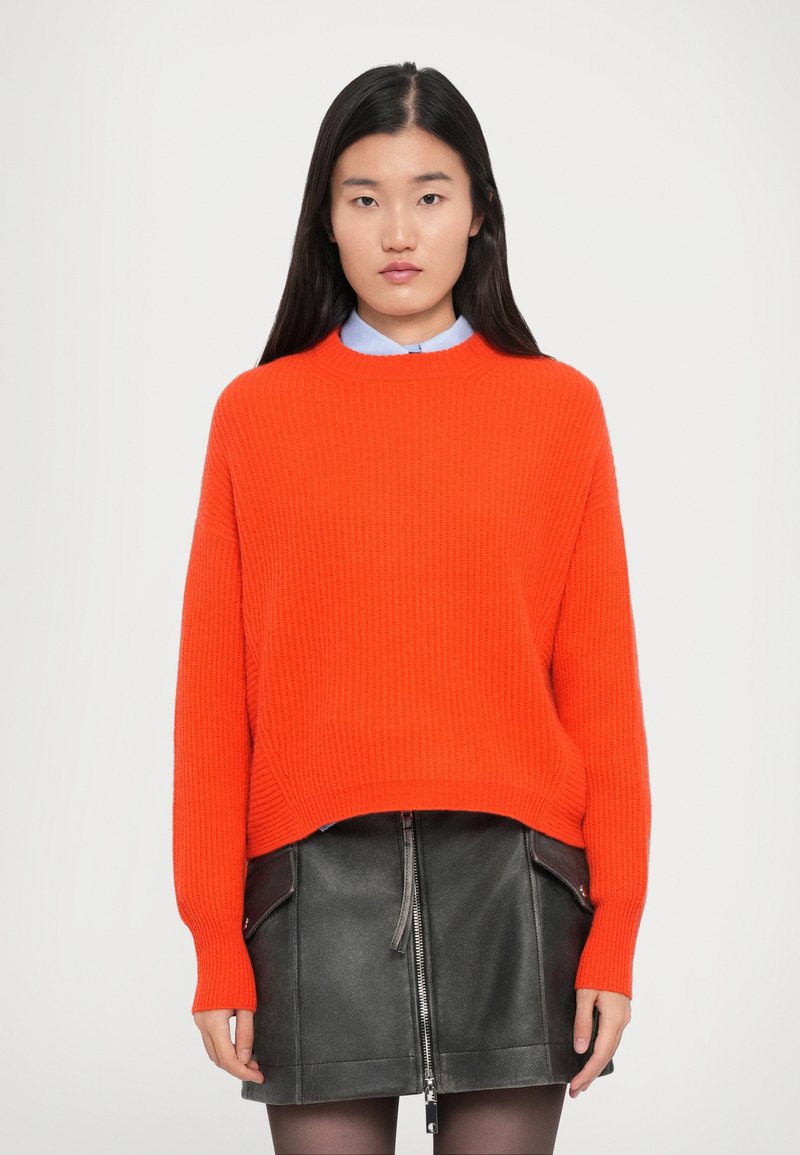 Maglione a coste di un brillante arancione con maniche lunghe, abbinato a una gonna in pelle nera con zip centrale e tasche laterali, indossato sopra una camicia azzurro chiaro.
