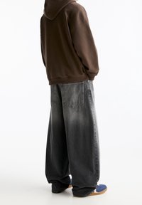 Sweat à capuche marron, pantalon en denim gris surdimensionné avec un effet délavé, grandes poches et ouverture large aux jambes. Porté avec des sandales bleues à glissière.