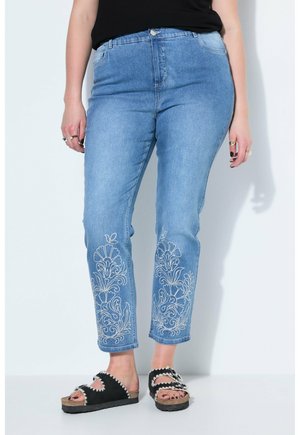 Kvinde iført lyseblå jeans med hvid blomsterbroderi på underbenene og sorte slip-on sandaler med korksål.