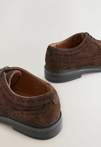 Brogues en daim marron avec des détails perforés, doublure en cuir et semelle en caoutchouc noir. Présente une texture lisse et un design classique.
