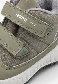 Zielone sportowe buty z tkaniny z rzepami, teksturowanym cholewką, amortyzowanym kołnierzem i kontrastującą białą gumową podeszwą z wzorzystym bieżnikiem.