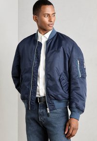 Mørkeblå bomberjakke med glidelås, ribbestrikkede mansjetter og nederkant, sidelommer og en kontrastlomme på venstre erme.