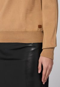 Beige Strickpullover mit Rippstrickbündchen und Saum, versehen mit einem kleinen braunen Leder-Logo-Tag in der Nähe der unteren Naht, kombiniert mit einem schwarzen Lederrock.