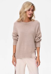 Beżowy sweter z dzianiny o szerokim dekolcie, z prążkowanymi mankietami i dołem. Oferuje luźny krój z subtelnymi bocznymi rozcięciami. Gładka tekstura.