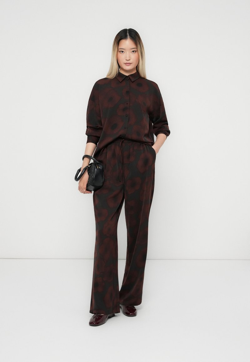 Bruine bloemenpatroon bijpassende shirt en broek, gemaakt van een gladde stof. Model houdt een kleine zwarte handtas vast en draagt donkere schoenen.