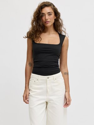 Femme aux cheveux bouclés portant un haut noir sans manches à encolure carrée et un pantalon blanc taille haute sur un fond clair uni.