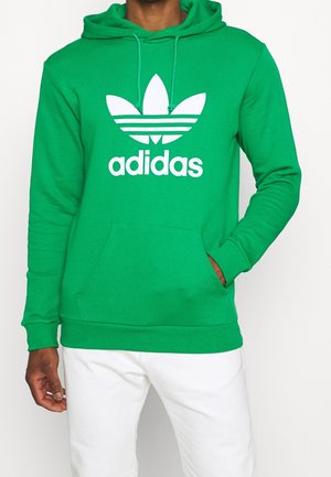 Mężczyzna ubrany w zieloną bluzę z kapturem Adidas z białym logo i napisem, w białych spodniach, stoi z jedną ręką w kieszeni.