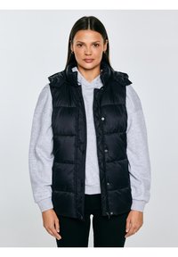 Naine kannab musta puffervesti helehallide kapuutsiga ja mustade pükstega, seistes ühevärvilise hele tausta ees.