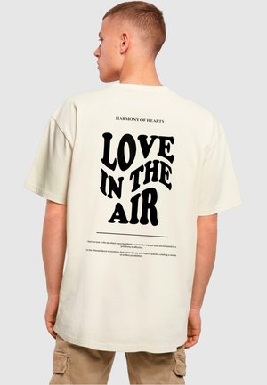T-shirt surdimensionné couleur crème avec du texte noir en gros sur le dos : "L'AMOUR DANS L'AIR" et un texte plus petit au-dessus dans une police moderne. Texture de tissu naturel.