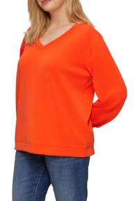Orange V-Ausschnitt Sweatshirt aus weichem Stoff in lockerer Passform, mit langen Ärmeln und gestickten Saumdetails, kombiniert mit blauen Jeans.