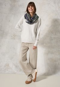 Lysegrå sweater med ribbet kant, beige bukser med bred benvidde og cremefarvede sneakers med brune såler. Modellen bærer et mønstret sort og grønt tørklæde.
