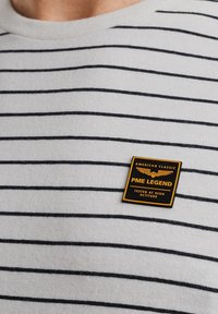 Chemise grise à rayures noires, avec un patch carré jaune et noir portant l'inscription "PME LEGEND" sur le côté gauche.