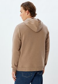 Sudadera de algodón marrón con capucha ajustable, puños acanalados y dobladillo. Combinada con jeans de mezclilla azul, mostrando una textura lisa y sencilla.
