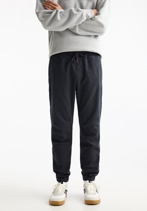 Person wearing black jogger pants with elastic cuffs, a grey sweatshirt, and white sneakers with gum soles, standing with arms crossed.