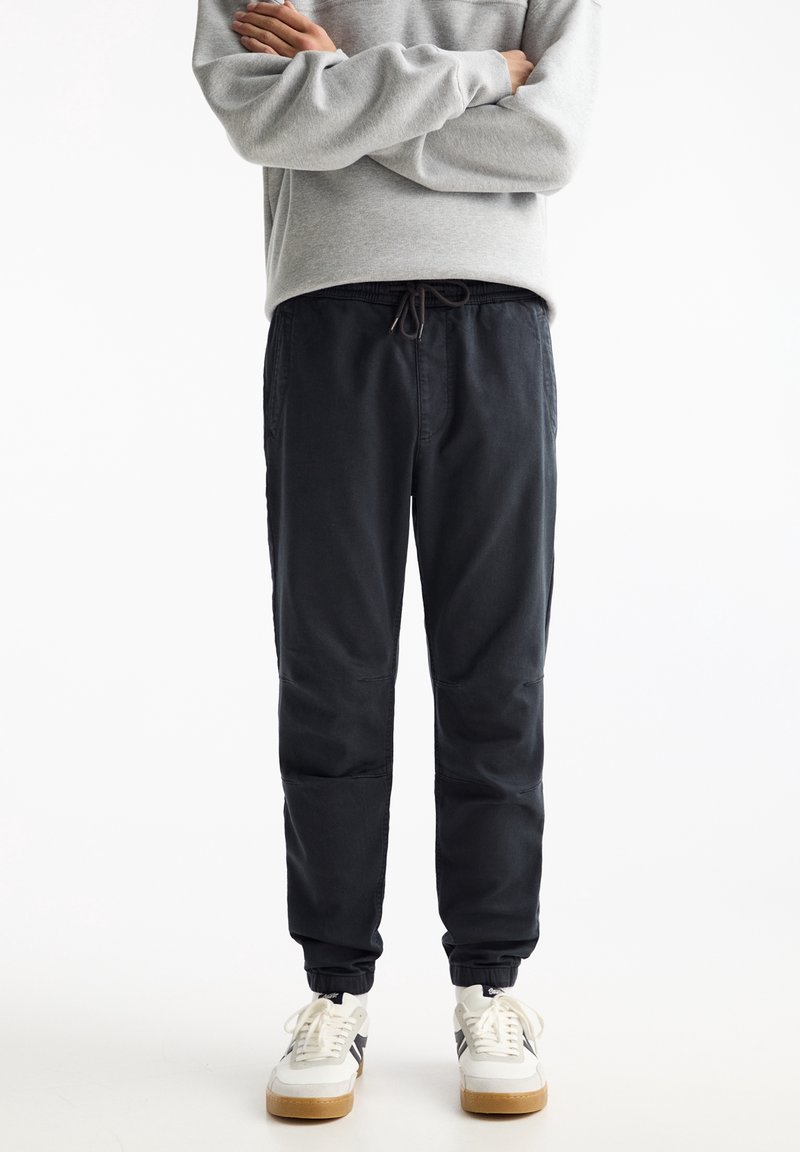 Personne portant un pantalon de jogging noir avec des poignets élastiques, un sweat-shirt gris et des baskets blanches à semelles en gomme, debout les bras croisés.