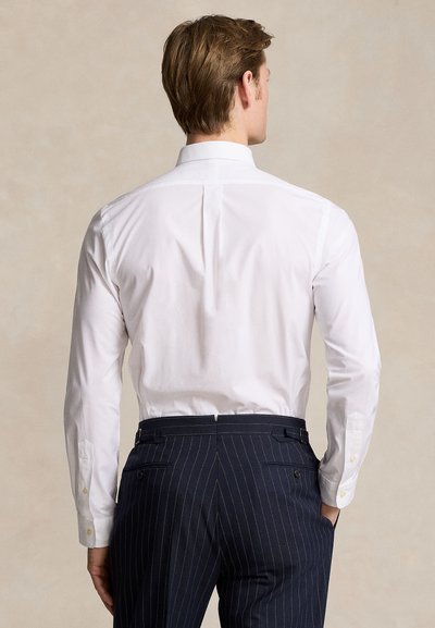 Muž v bielej košeli s dlhým rukávom a tmavomodrých nohaviciach s prúžkami stojaci s rukami vreckách, otočený chrbtom.