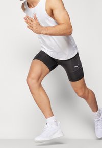 Pantalones cortos deportivos negros con el logo de Puma, combinados con una camiseta sin mangas blanca y zapatillas blancas. El modelo está en una pose de carrera.