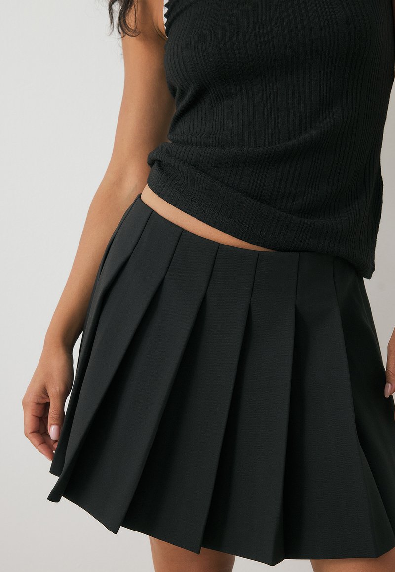 Femme portant un haut sans manches en maille noire et une mini-jupe plissée noire taille haute, se tenant devant un fond clair uni.
