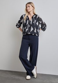 Schwarz-weiß gemusterte Bluse mit geometrischem Muster und langen Ärmeln, kombiniert mit navyblauen weiten Hosen und weißen Sportschuhen.