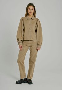 Chaqueta de tela texturizada de color marrón claro, con cuello y diseño de mangas abullonadas, que presenta botones plateados. Combinada con pantalones a juego y zapatos negros.