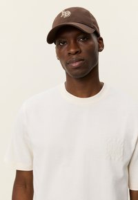 Καφέ καπέλο μπέιζμπολ με λογότυπο, συνδυασμένο με λευκό κοντομάνικο μπλουζάκι που έχει μικρό υφασμάτινο μπάλωμα στο αριστερό στήθος. Απλός σχεδιασμός.