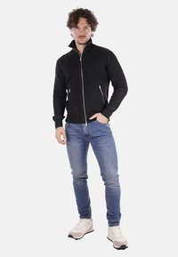 Giacca nera a coste con zip e colletto alto, abbinata a jeans blu e sneakers chiare. Tessuto liscio, design attillato e dettagli in metallo argentato.