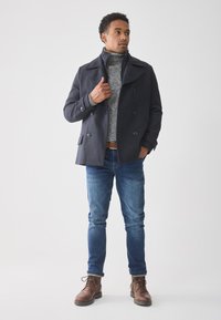 Abrigo marino de doble botonadura sobre un cuello de tortuga gris, combinado con jeans azules y botas marrones de cordones. Tejido suave, corte entallado.