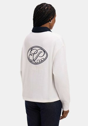 Personne aux cheveux tressés portant un haut blanc à manches longues avec un col sombre et un logo d'initiales brodé dans le dos, assorti à un jean foncé.