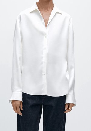 Person iført en hvid langærmet skjorte med knapper og krave samt mørkeblå jeans, stående foran en ensfarvet lys baggrund.