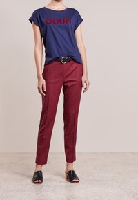 T-shirt bleu marine avec texte "HUGO" rouge, associé à un pantalon fuselé à carreaux rouges et à des mules noires à bout ouvert. Tissu texturé, coupe décontractée.