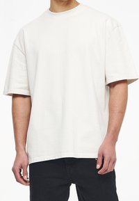 Světle béžové bavlněné tričko s krátkým rukávem, klasickým kulatým výstřihem a decentním logem na přední straně. Nosené s tmavými džínami.