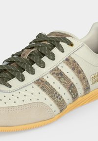 Baskets blanches Adidas avec trois bandes à motifs, lacets vert olive, bout en daim beige et semelle en caoutchouc.