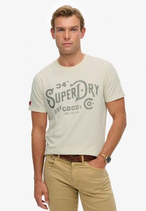 Licht beige T-shirt met een grote grijze grafische "SuperDry"-bedrukking; ronde hals, korte mouwen, gedragen met kaki broek en een bruine gevlochten riem.