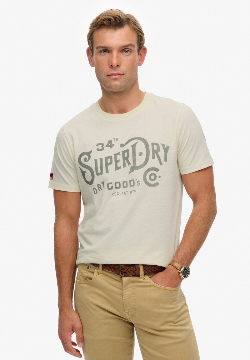 Camiseta de color beige claro con un gran estampado gráfico gris de "SuperDry"; cuello redondo, mangas cortas, combinada con pantalones caqui y un cinturón marrón tejido.