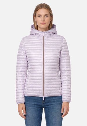 Kobieta stojąca przodem, ubrana w jasnofioletową pikowaną kurtkę z kapturem i przednim zamkiem błyskawicznym oraz niebieskie dżinsy na jednolitym tle.