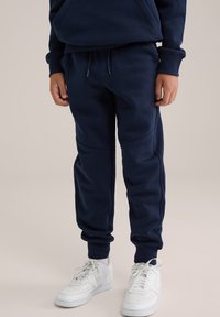 Jūras zila, mīksta materiāla sporta bikses ar brīvu piegriezumu, elastīgu jostu ar auklu un ribotām aprocēm. Apvienotas ar baltām sporta kedām.