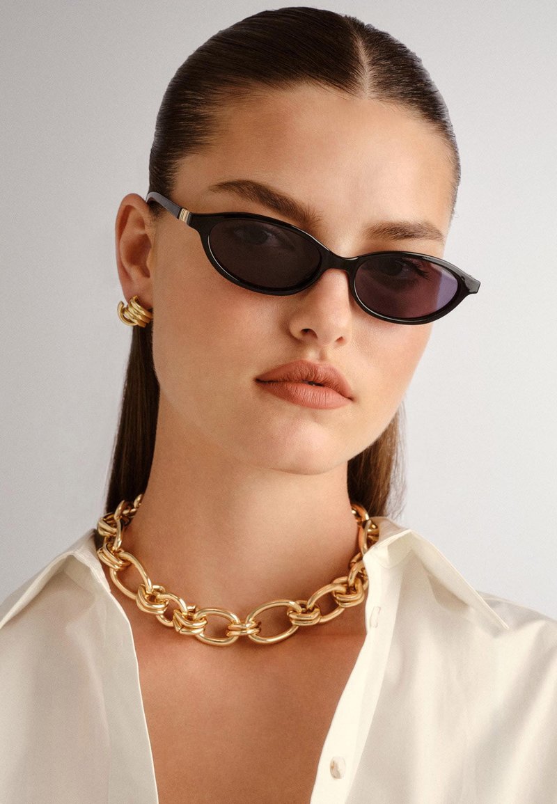 Black oval sunglasses with dark lenses, gold chain necklace, gold hoop earrings, and a white collared shirt. Smooth textures and modern design.