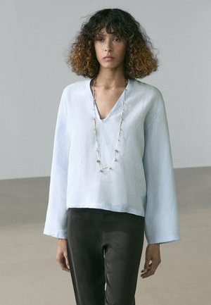 Femme aux cheveux bouclés portant un chemisier ample bleu clair à col en V, un long collier et un pantalon noir, debout devant un fond gris uni.