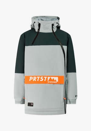 Persoon die een groene en grijze skikleding draagt met een oranje logo op de jas, een zwarte muts, regenboogreflecterende zonnebril en zwarte laarzen, rechtop staand.