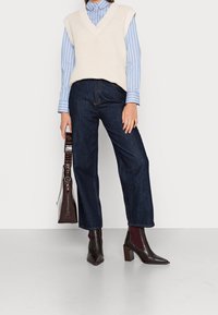 Crème gebreide vest over een blauw gestreept shirt, gecombineerd met donkere denim wijde broek en bruine haklaarzen, terwijl ze een bruine tas vasthoudt.