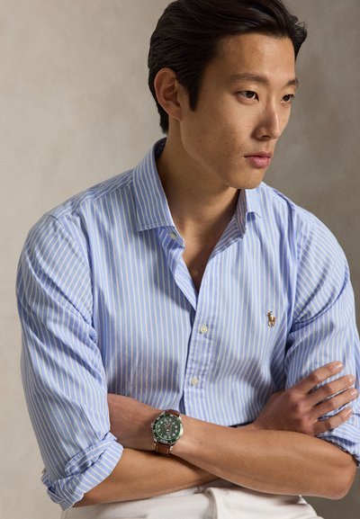 Mann trägt hellblau gestreiftes Hemd mit hochgekrempelten Ärmeln und eine grüne Armbanduhr mit braunem Lederarmband, Arme verschränkt, schaut nach rechts.